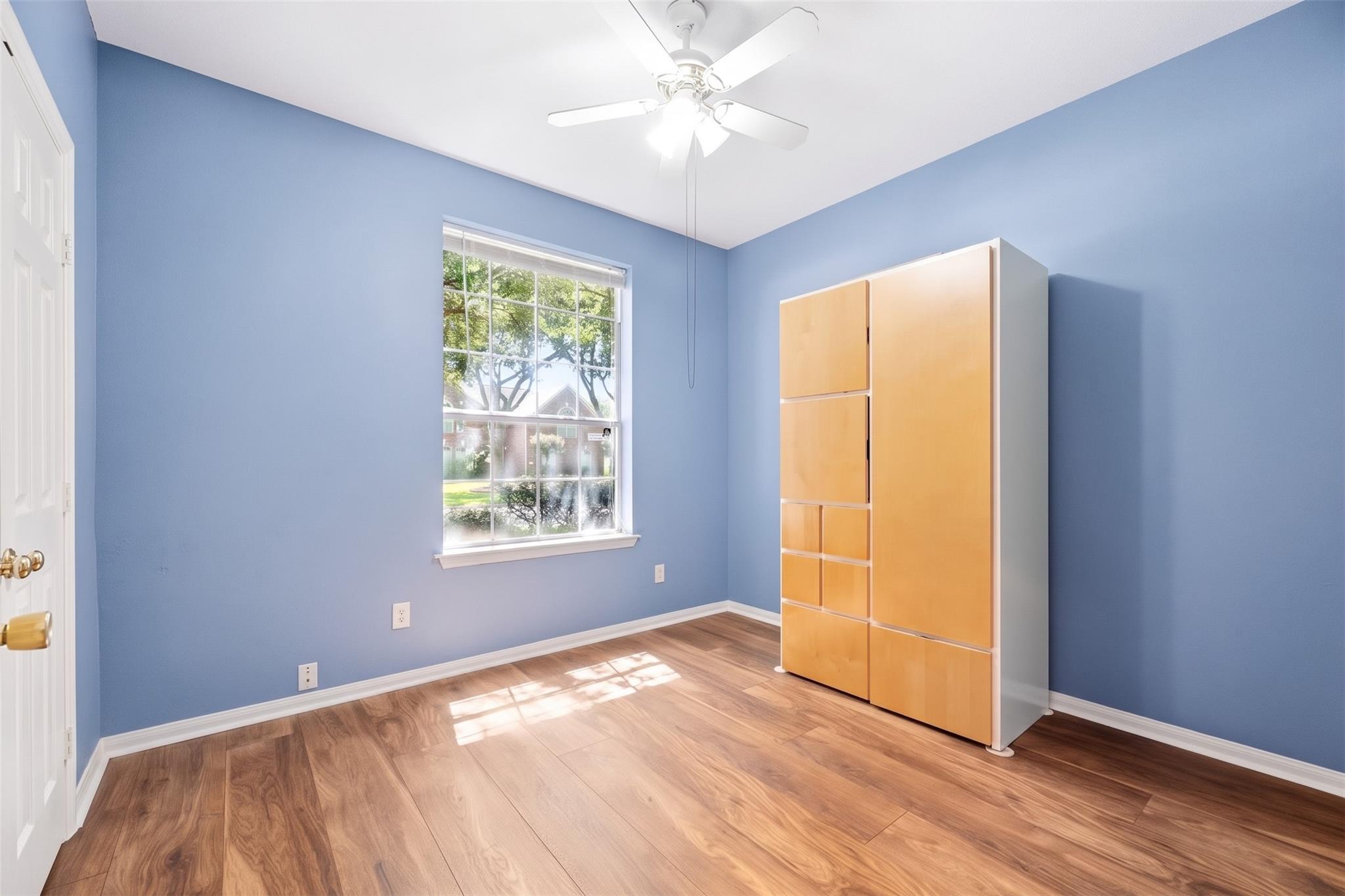 Blue-walled room with wood-look flooring, a large window, and a tall light-wood cabinet.