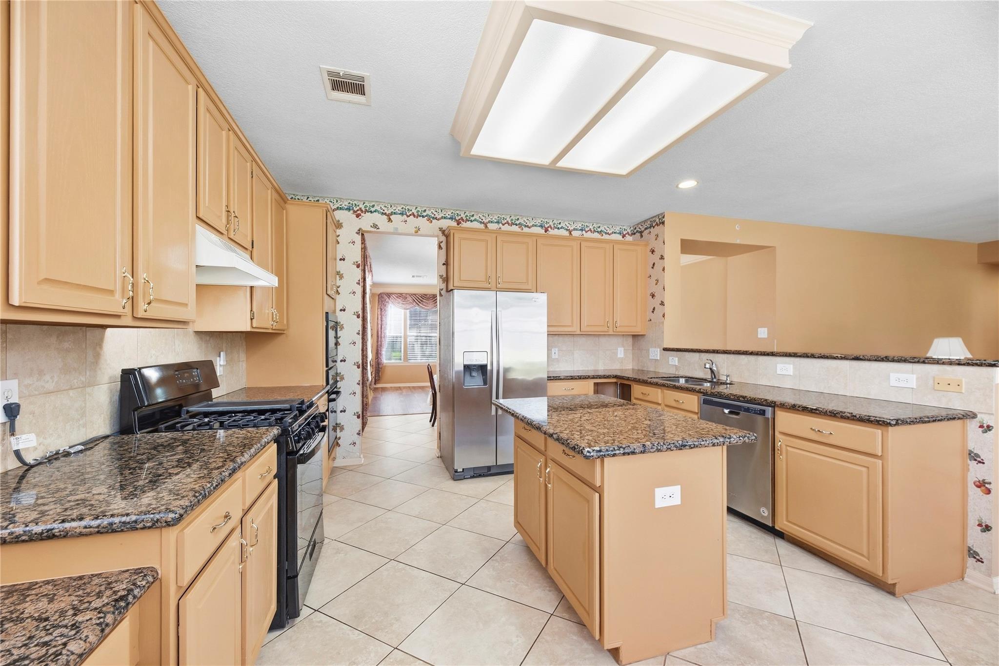 Kitchen with honey-colored cabinets, granite island, stainless steel appliances, and tiled floors.