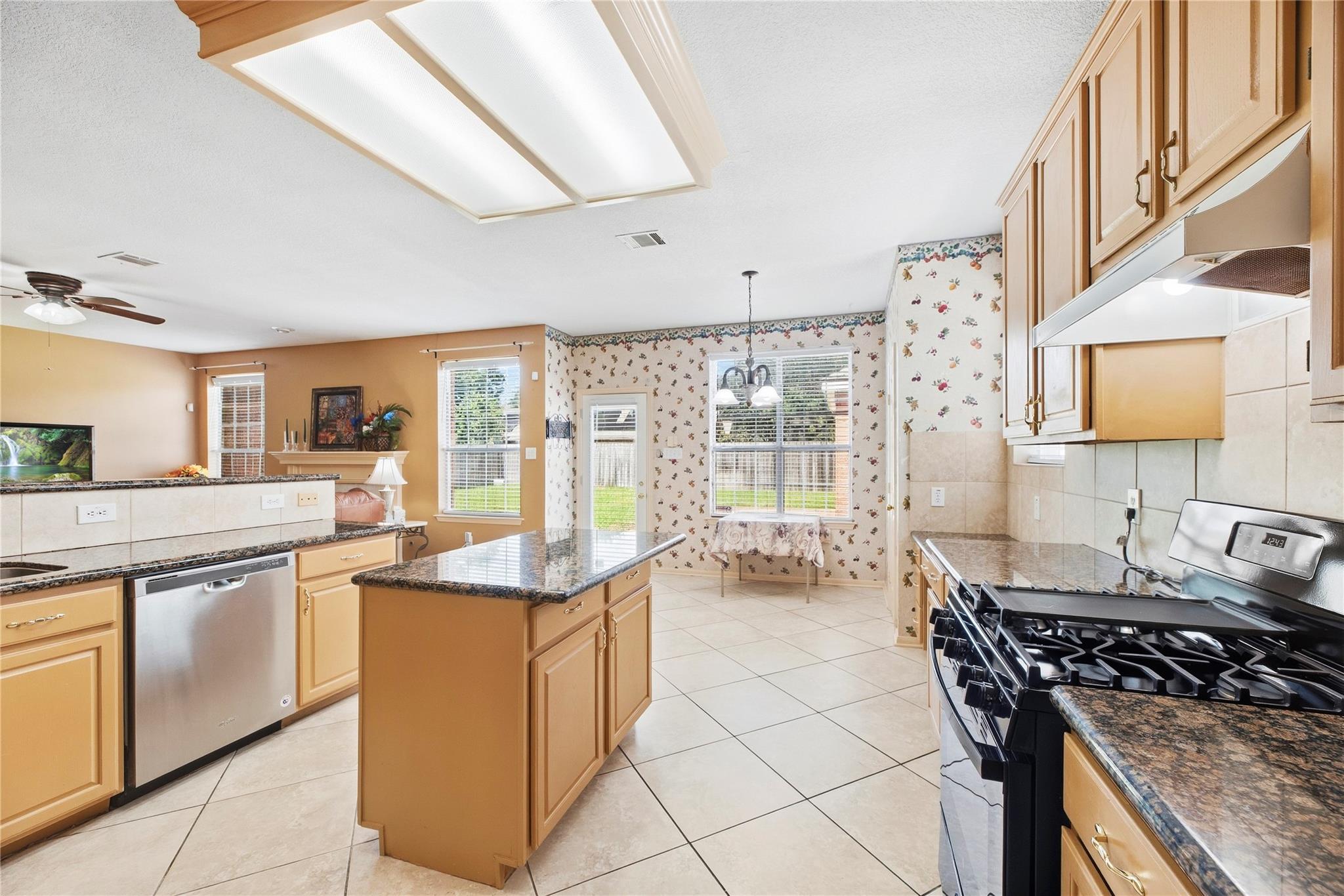 Kitchen with a central granite island, honey-colored cabinets, and floral wallpaper in the breakfast nook.