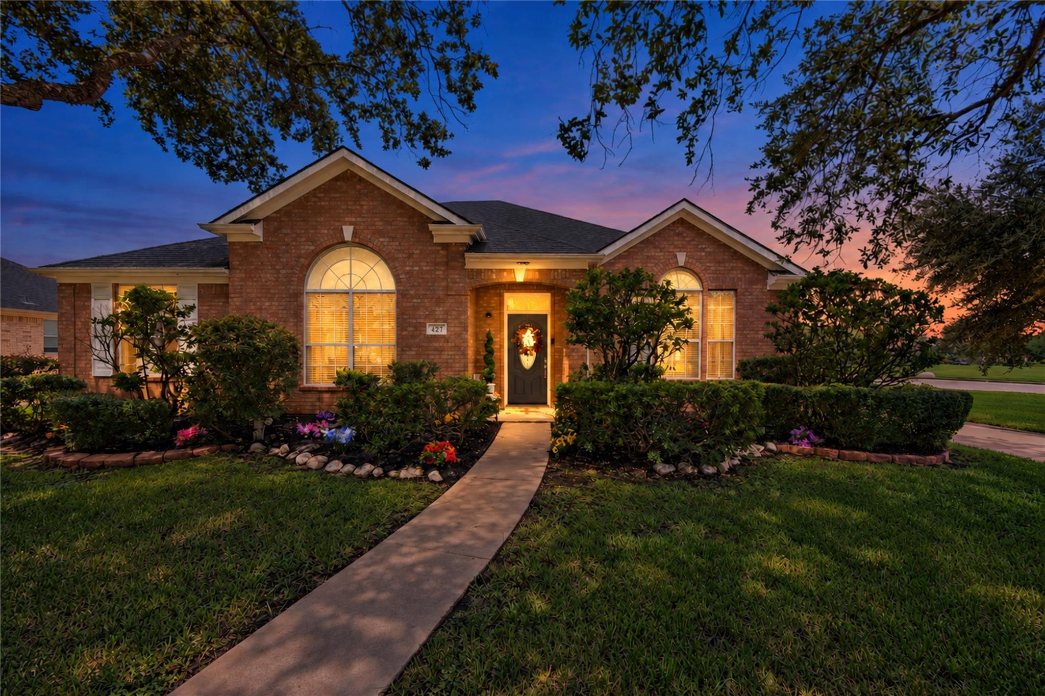 Red brick house at dusk with glowing arched windows and a curved walkway through landscaped gardens.