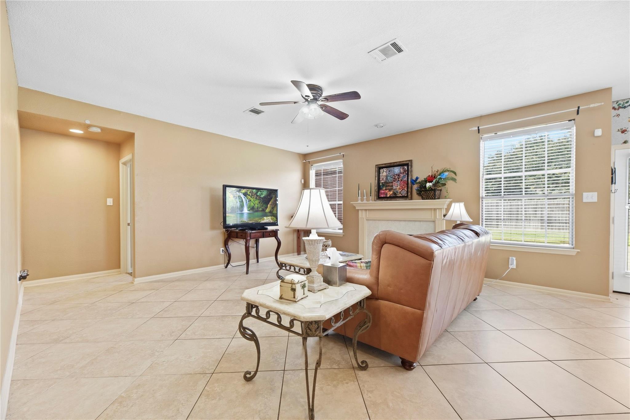 Furnished living room with a brown leather sofa, tiled floor, fireplace, and large windows with blinds.