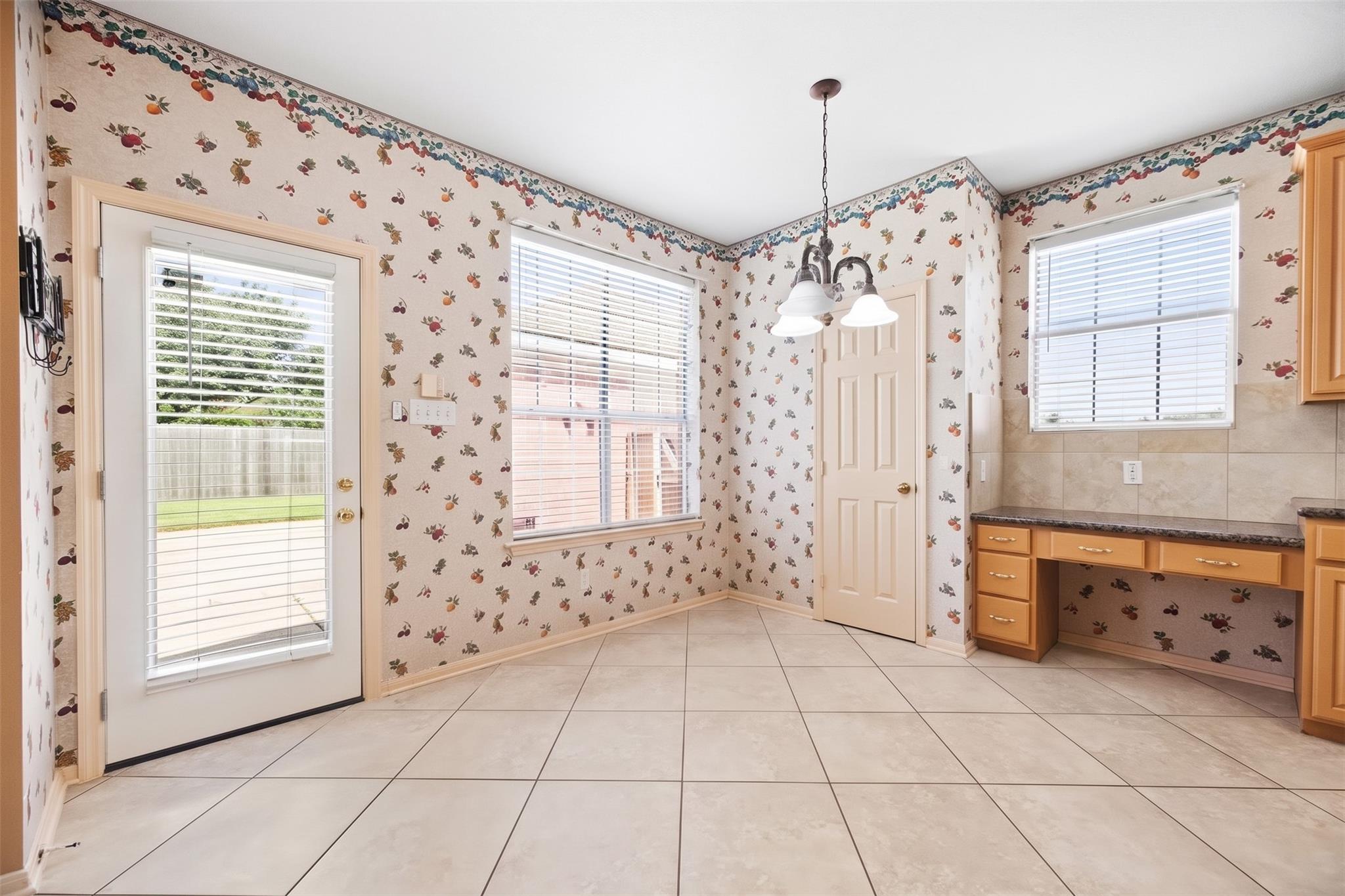 Unfurnished kitchen with fruit-patterned wallpaper, tiled floors, honey-colored cabinets, and windows with white blinds.