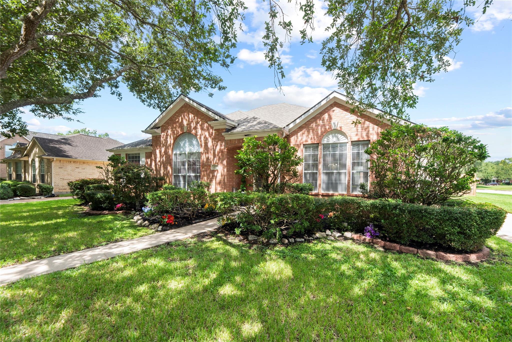 Red-brick house with arched windows, flowering garden beds, and a broad lawn beneath mature trees.