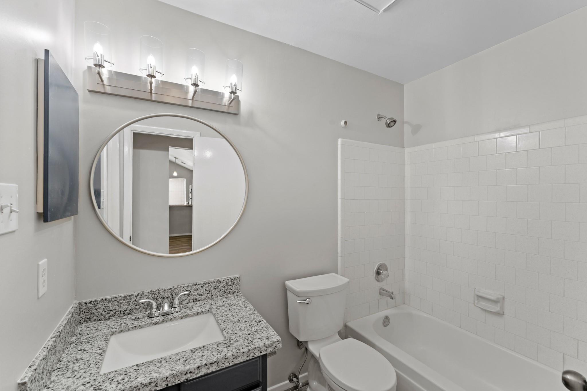 Bathroom with a granite vanity, round mirror, and white tiled tub and shower.