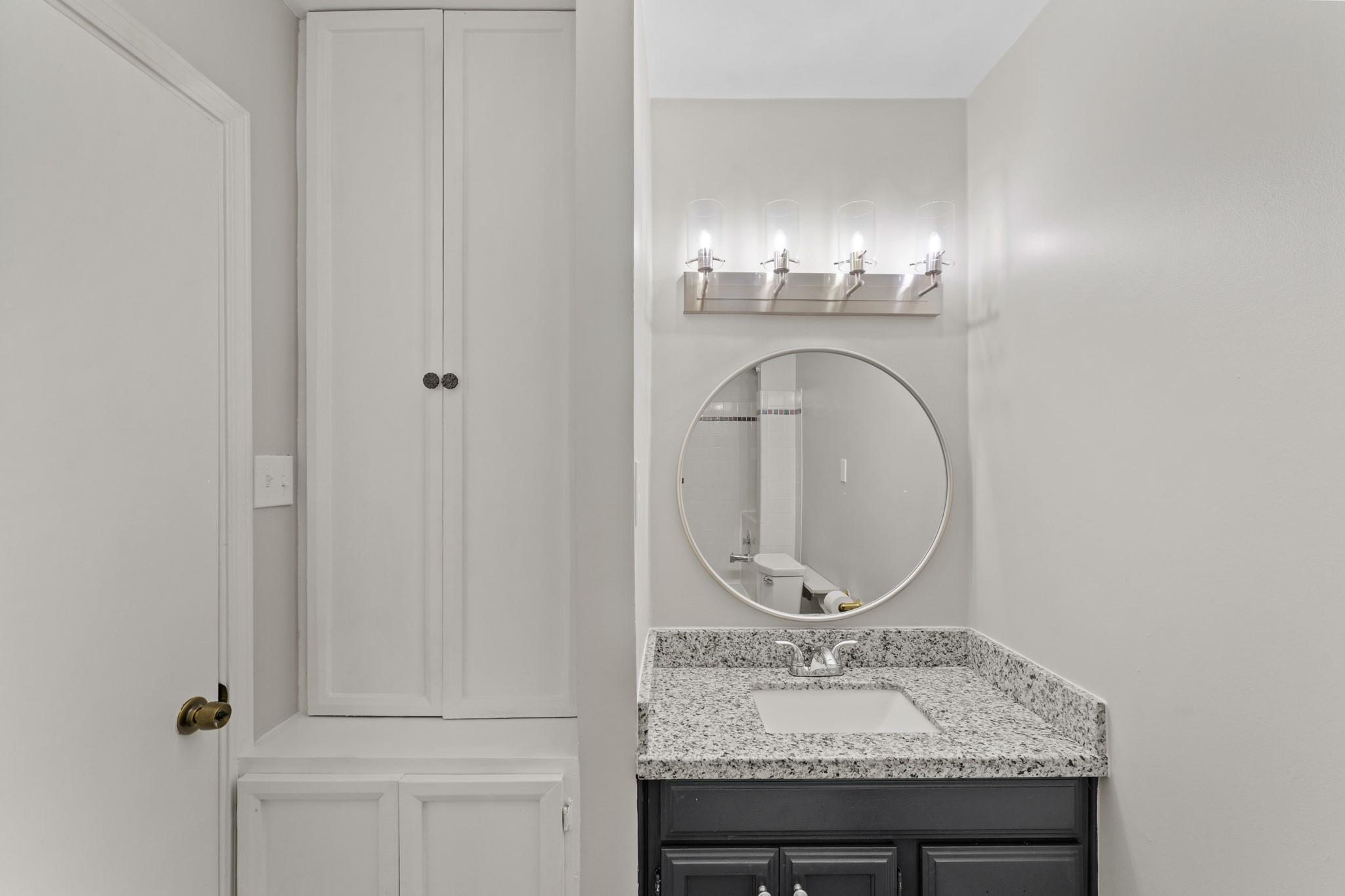 Bathroom vanity with speckled granite counter, dark cabinets, round mirror, and four-light fixture.