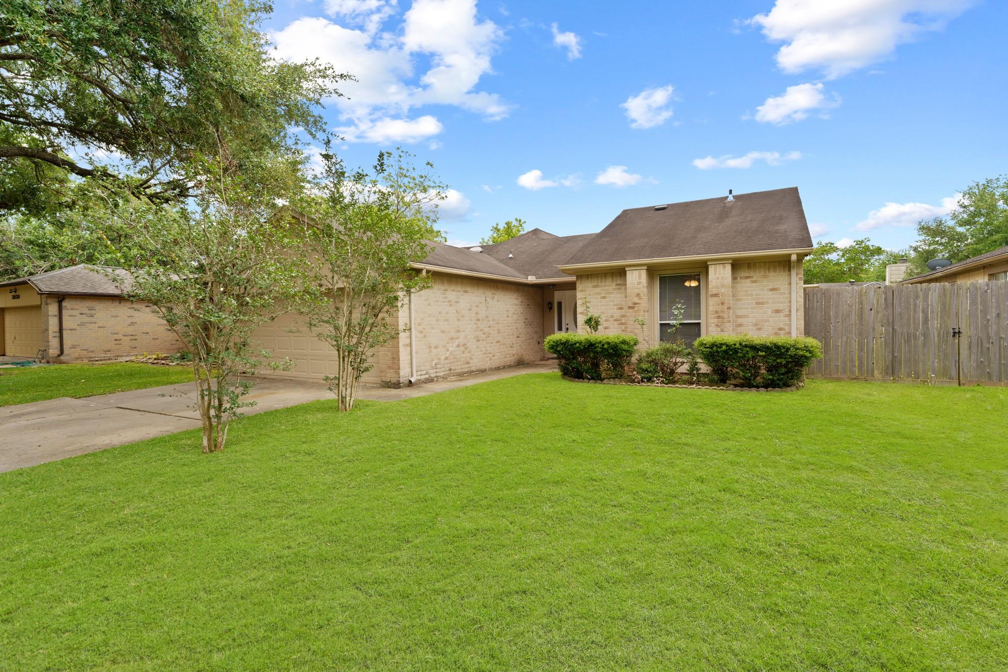 Tan brick home with an attached garage, broad green lawn, mature trees, and a wooden privacy fence.