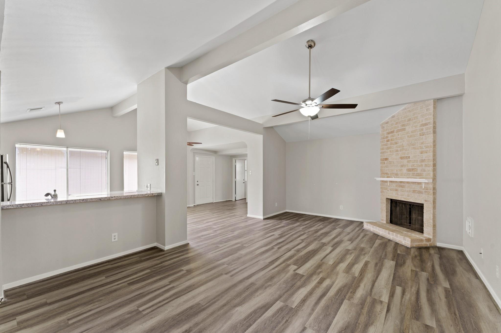 Vacant living room with vaulted ceiling, wood-look floors, and a light brick fireplace.