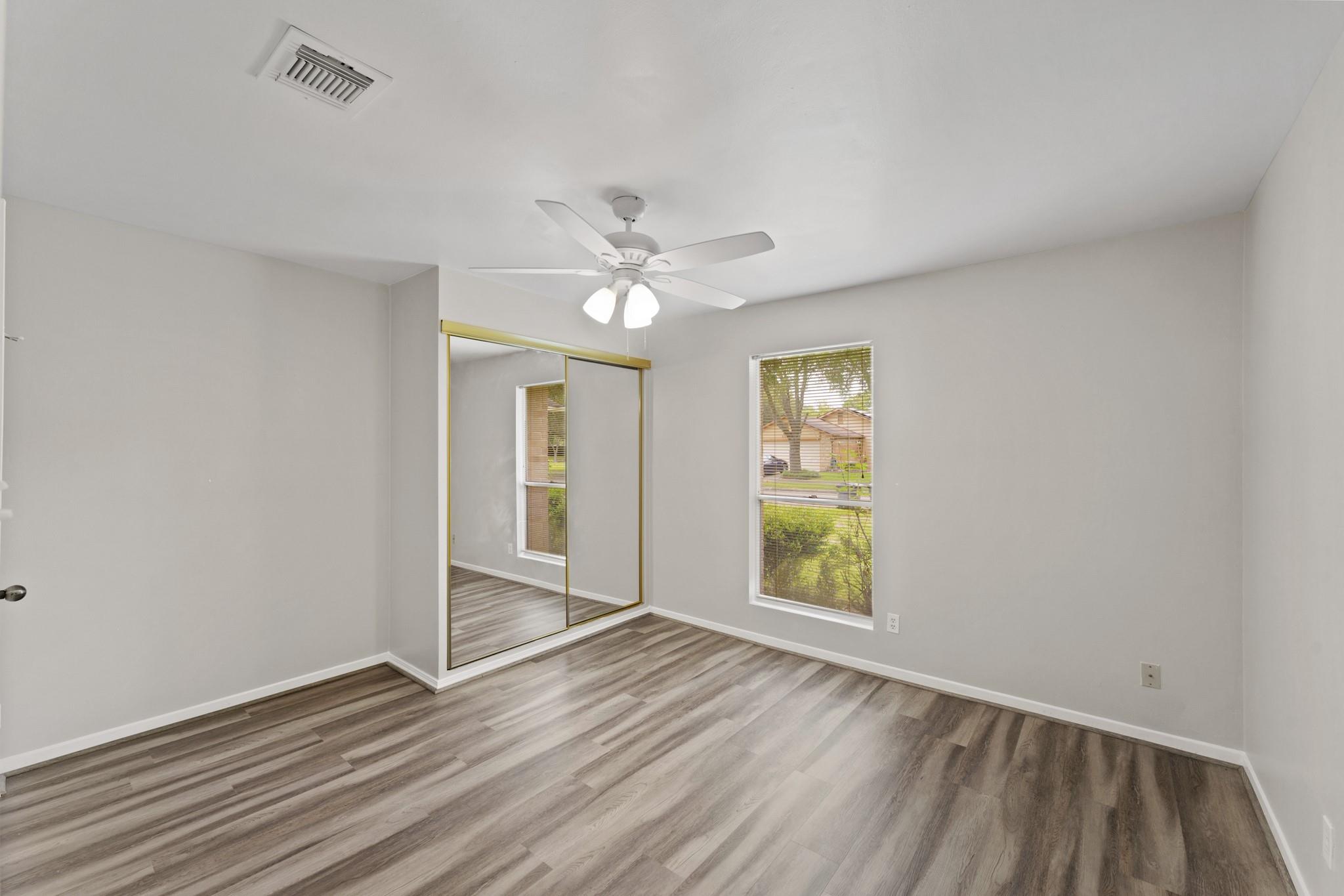 Empty bedroom with mirrored closet doors, wood-look flooring, and a ceiling fan.