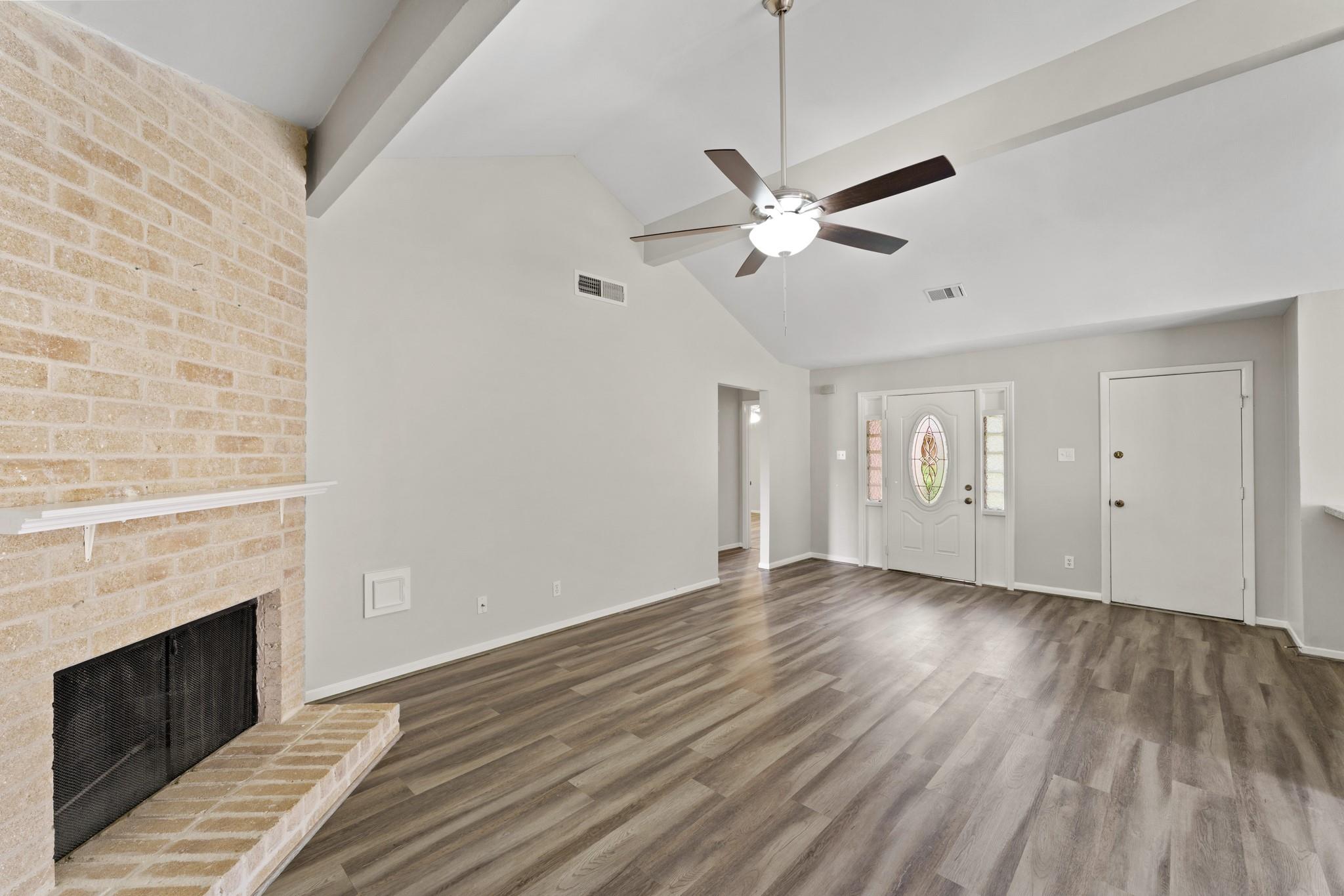 Empty living room with a tall brick fireplace, vaulted ceiling, and decorative glass front door.