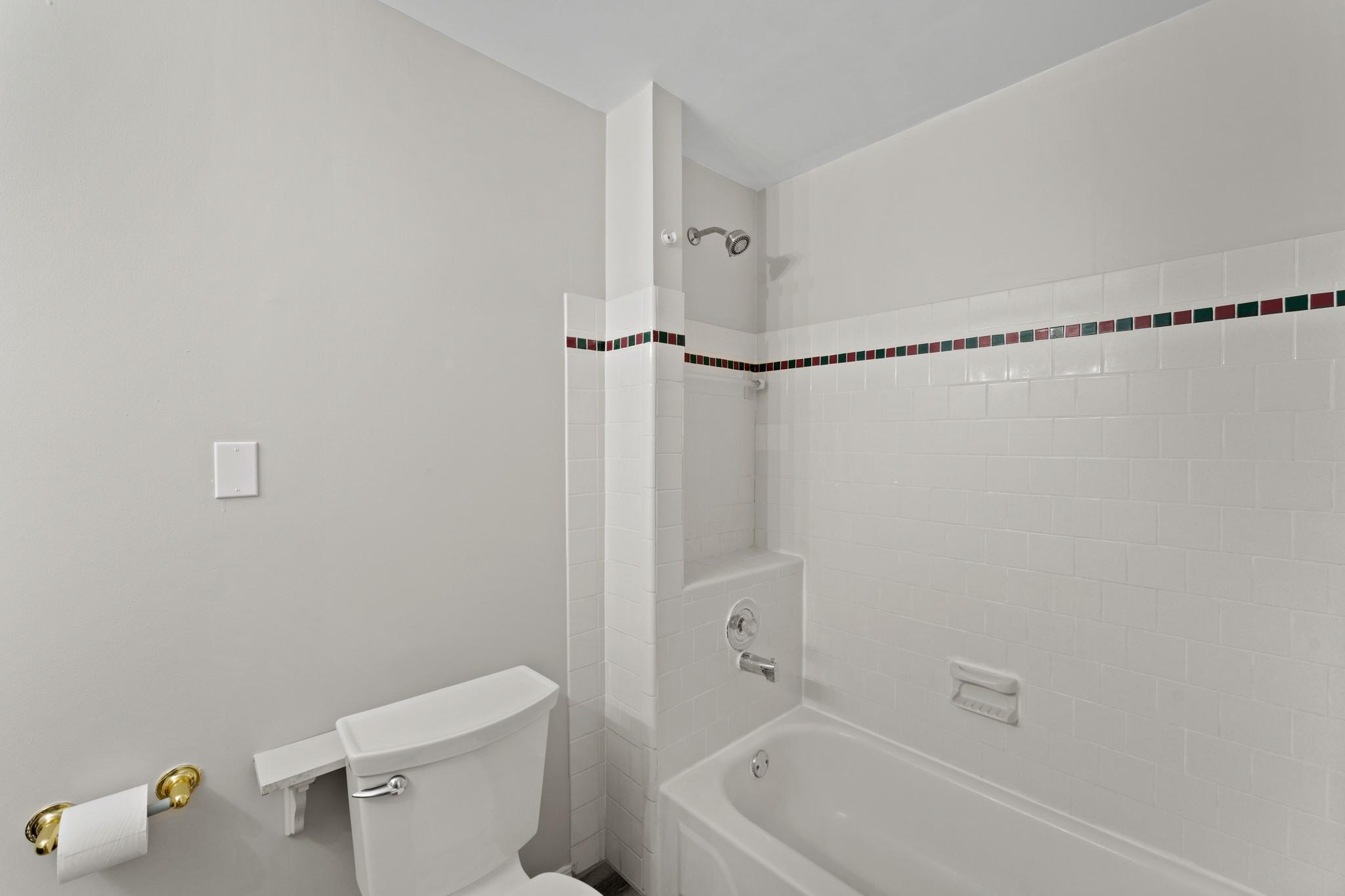 Bathroom with a white tub and shower, toilet, and red and green accent tile band.