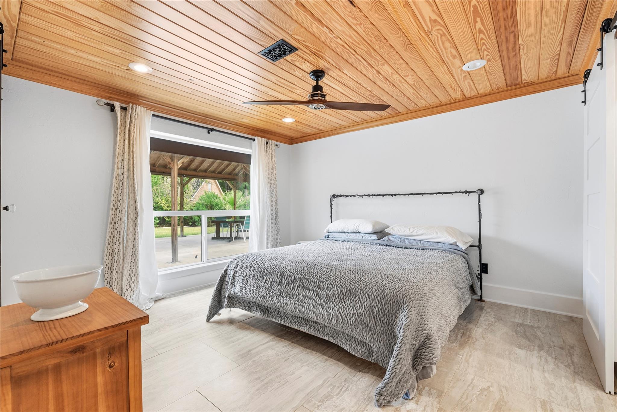 Bedroom with a wood plank ceiling, gray bed, and window overlooking a covered patio.