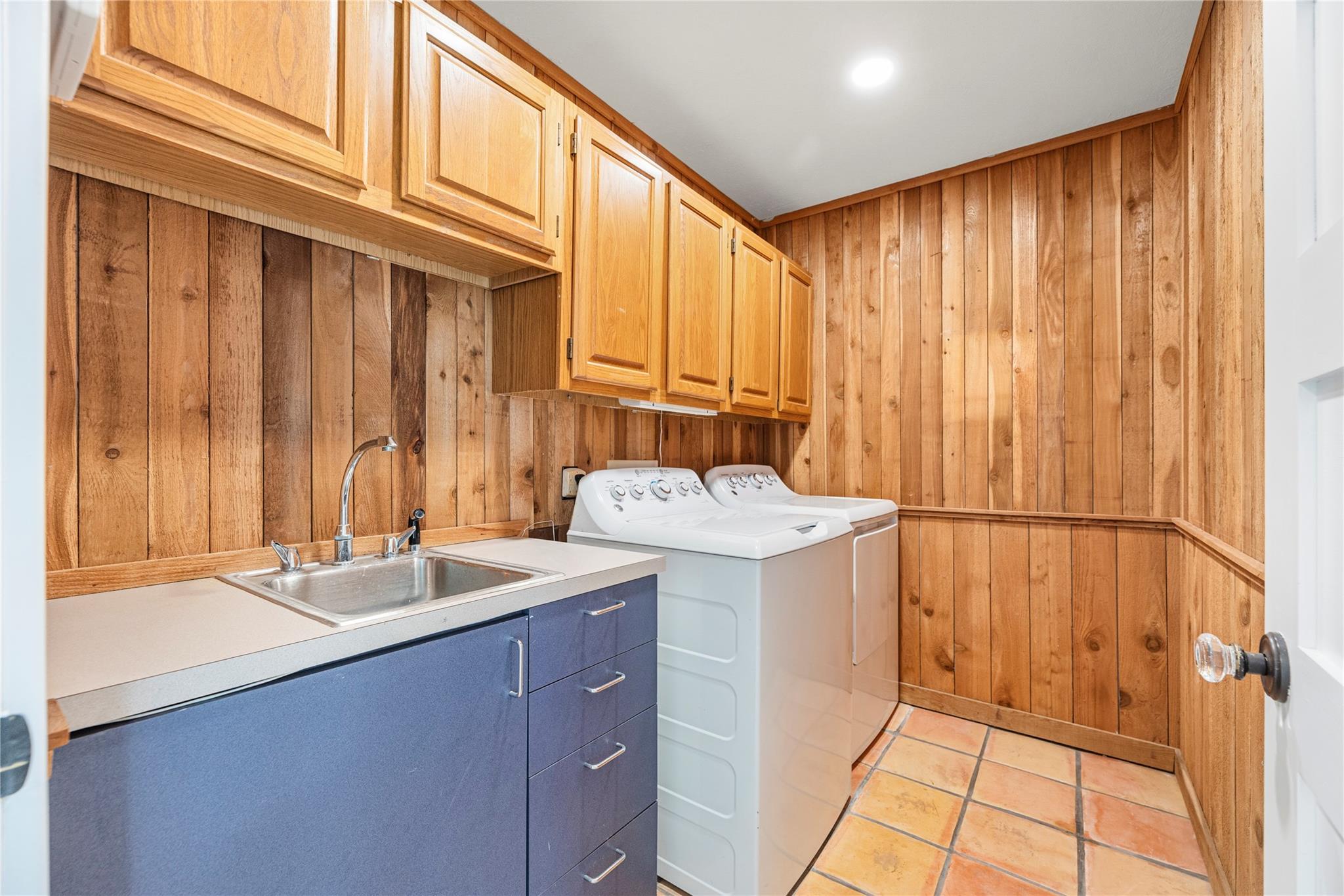 Laundry room with wood-paneled walls, overhead cabinets, washer and dryer, and a utility sink.