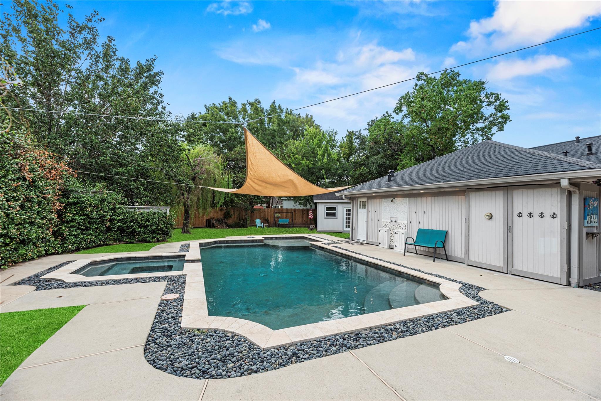 Backyard pool and raised spa beneath a tan shade sail, beside a white poolside outbuilding.