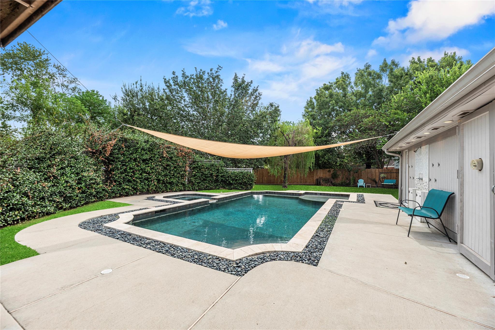 Backyard pool and raised spa beneath a shade sail, with stone edging and a concrete patio.