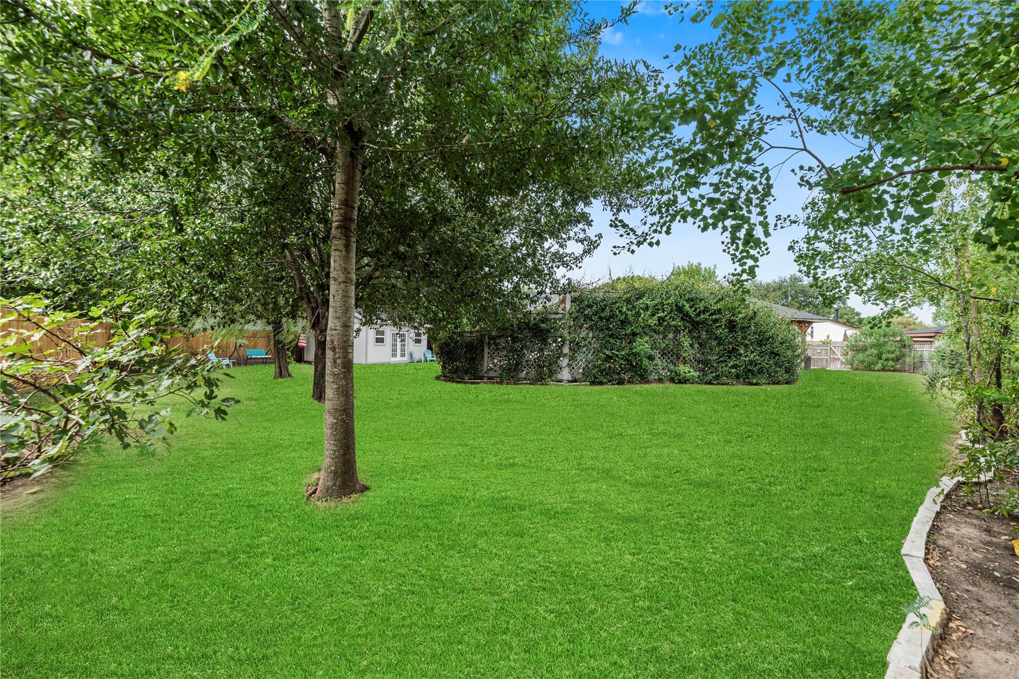 Backyard with a broad green lawn, mature trees, a tall hedge, and a small covered patio.