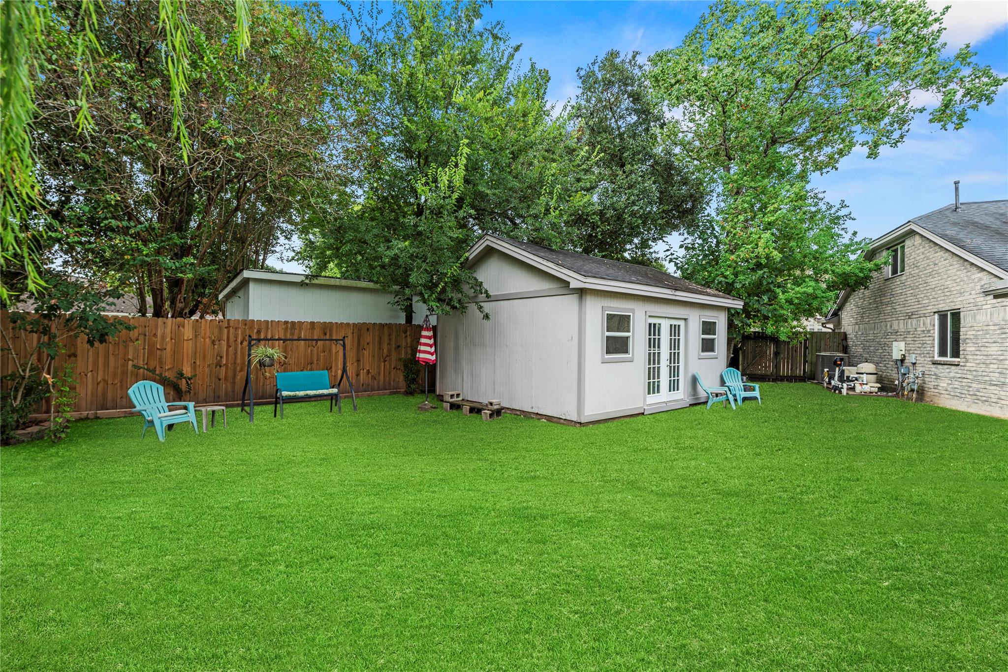 Backyard with a detached gray shed, green lawn, blue patio chairs, and mature trees.