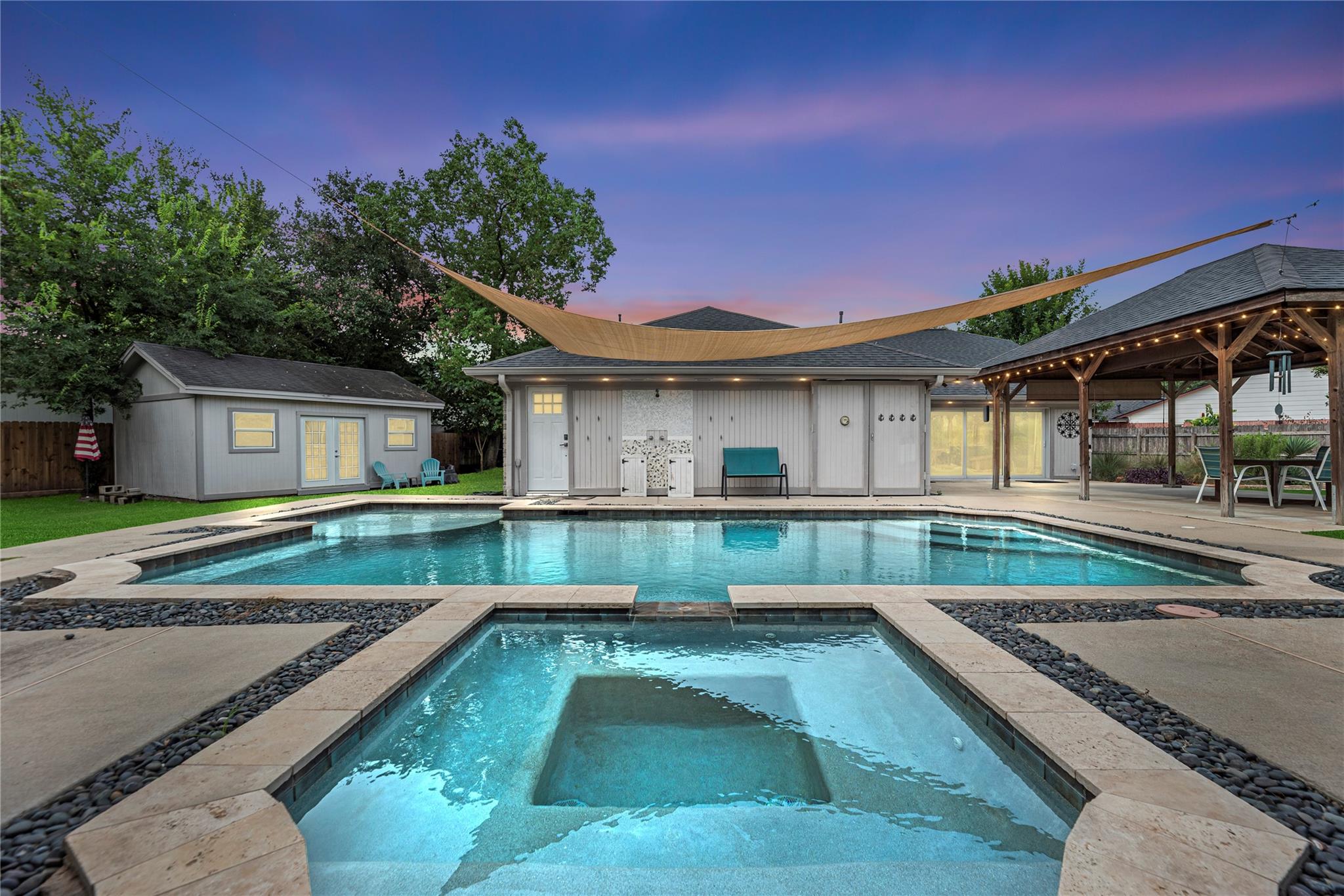 Backyard pool and raised spa beside a pool house and covered pavilion at dusk.