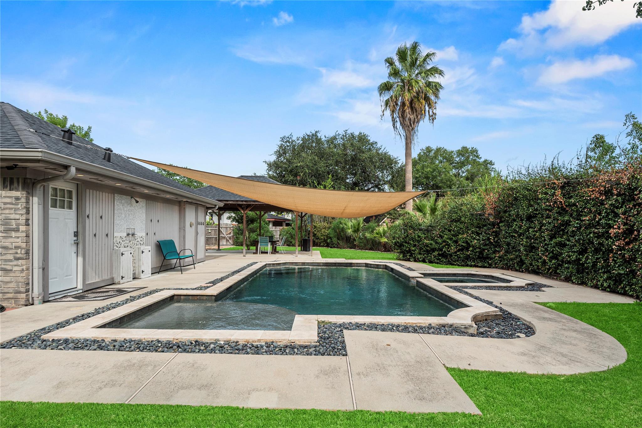 Backyard pool and spa beneath a tan shade sail beside the house.