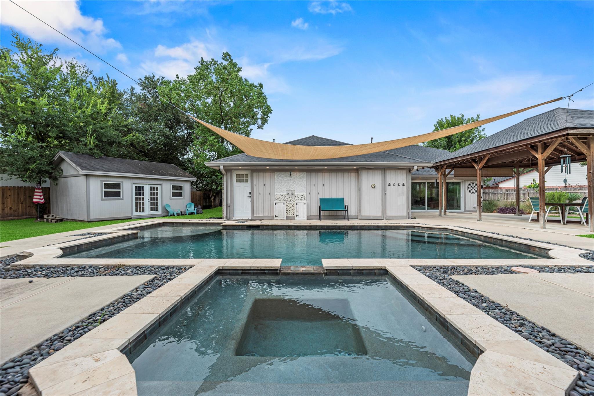 Backyard pool and spa facing a white pool house, with a shade sail and wooden pavilion.