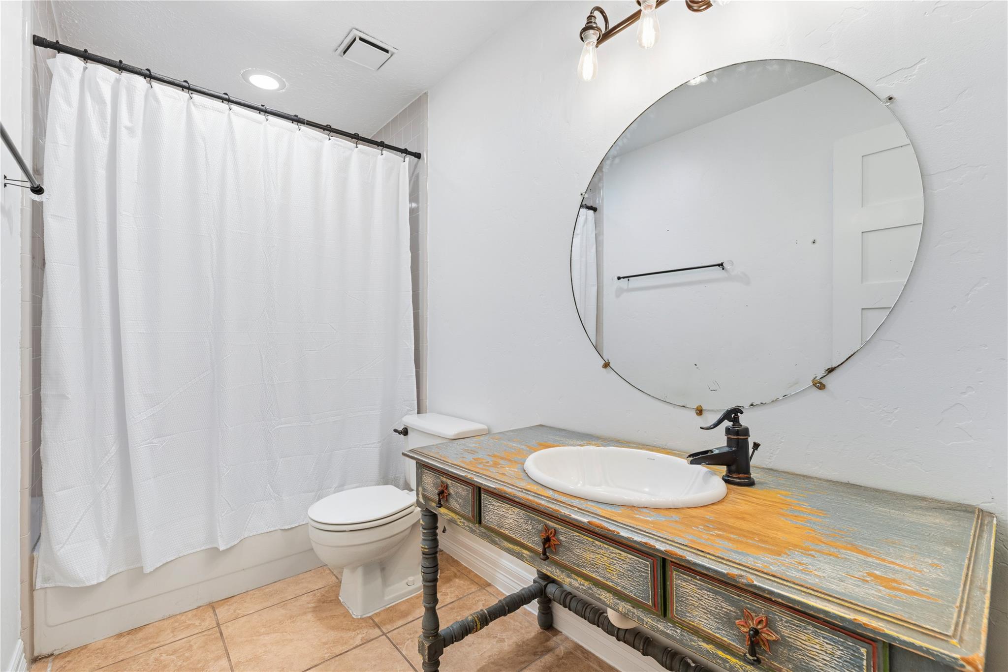 Bathroom with a distressed multicolored vanity, white sink, and oversized round mirror.