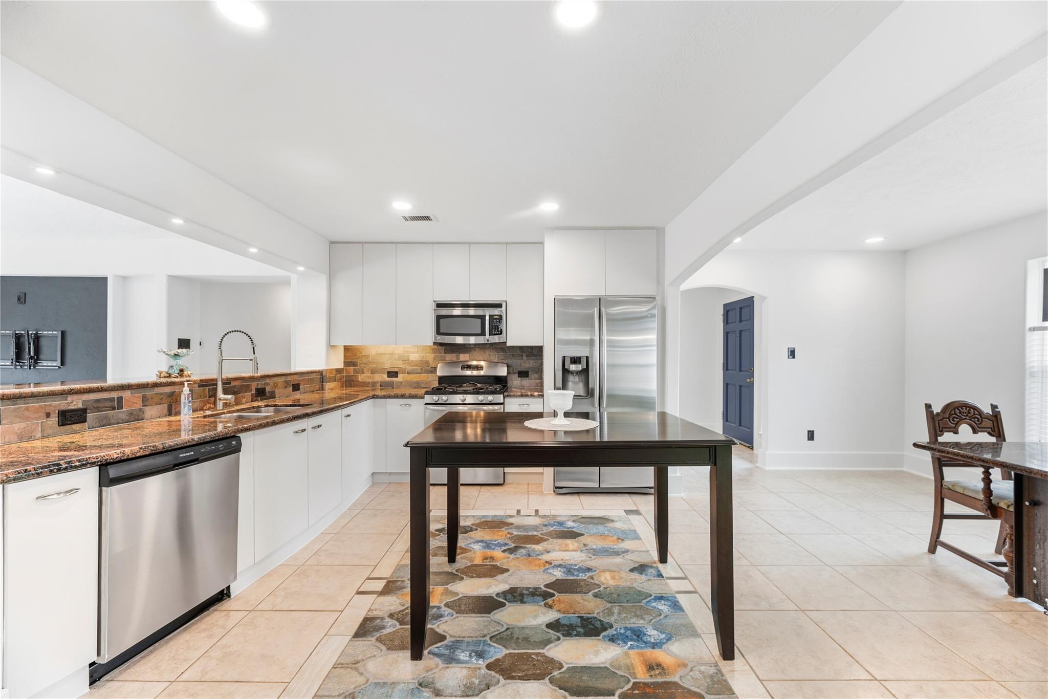 Kitchen with white cabinetry, stainless appliances, stone backsplash, and dark island table over a colorful tile runner.