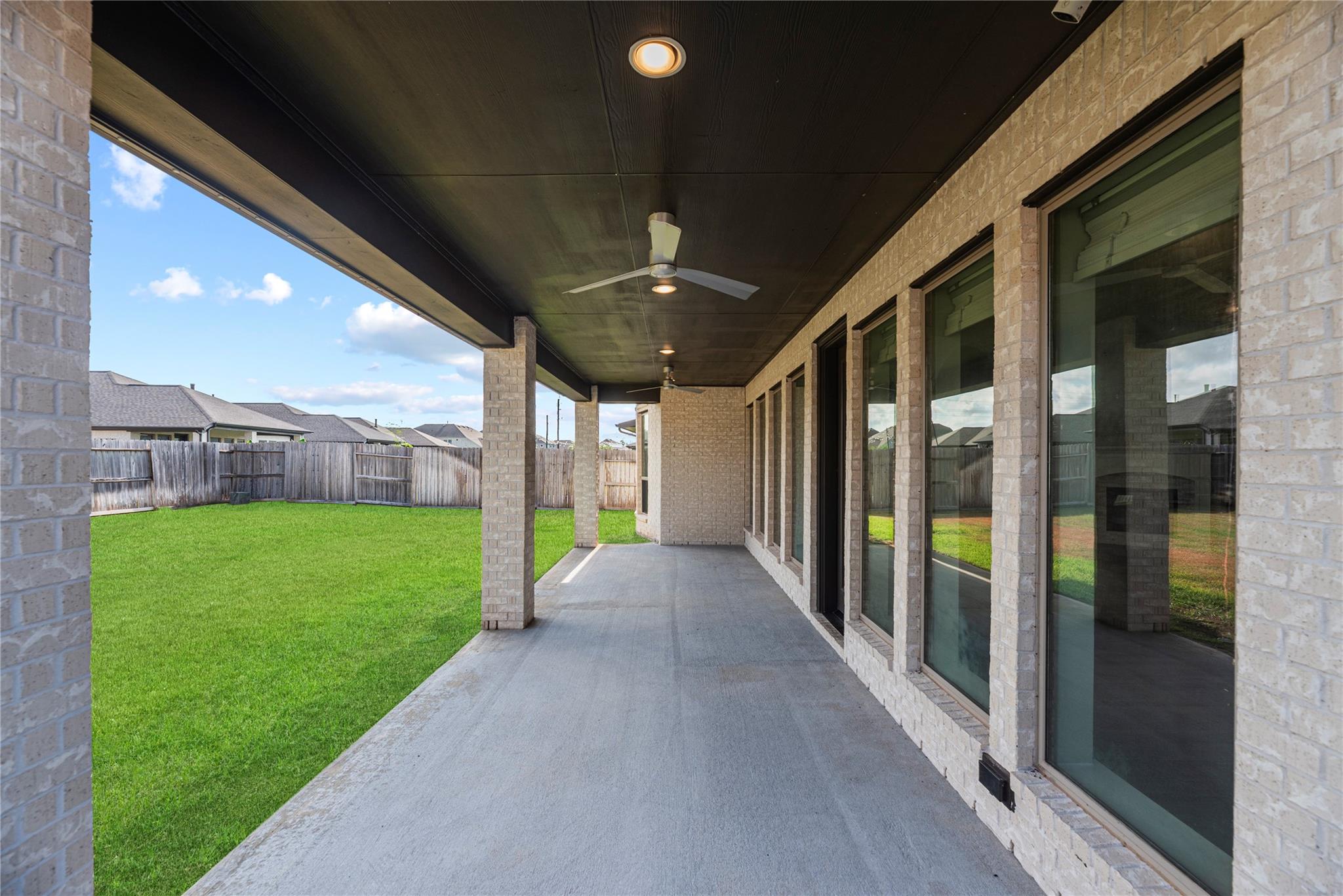Covered backyard patio with ceiling fans and a concrete floor overlooking a fenced lawn.