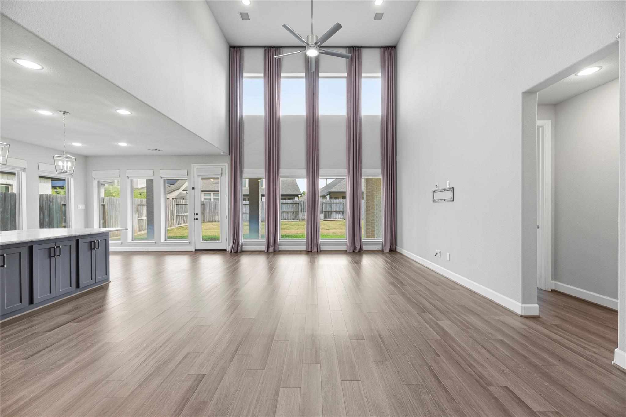 Vacant living area with double-height windows, mauve curtains, wood-look floors, and a kitchen island.
