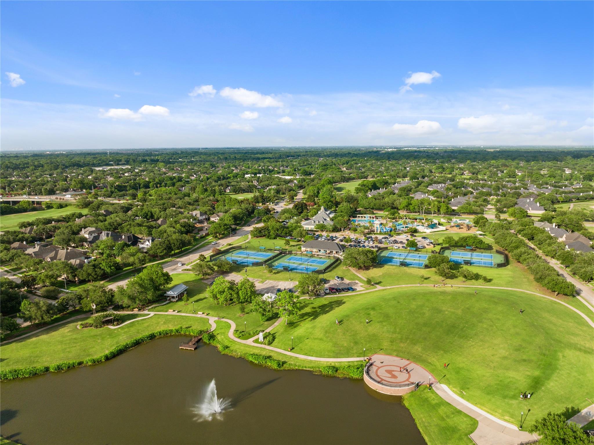 Aerial view of a community park with a pond, fountain, tennis courts, and pools.