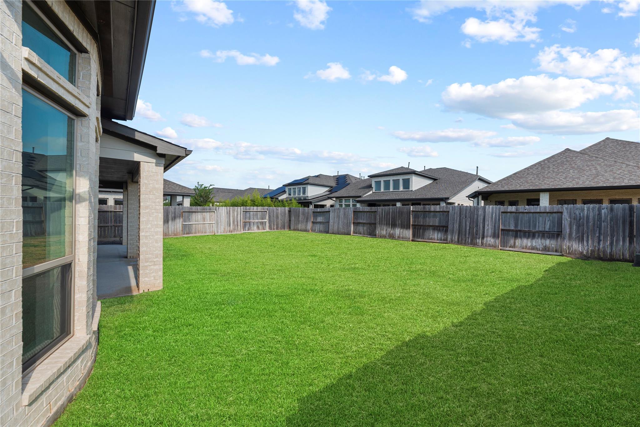 Fenced backyard with a wide green lawn beside a light-brick home and covered patio.