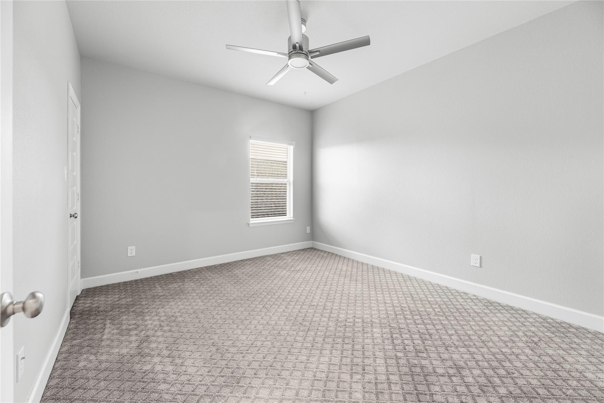 Empty bedroom with patterned gray carpet, a window with blinds, and a ceiling fan.