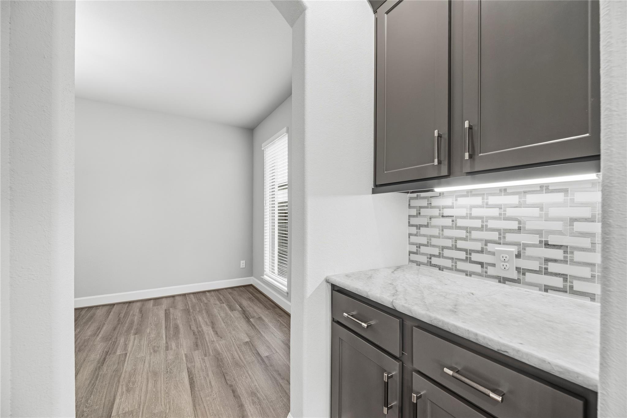 Kitchen counter with dark cabinets and patterned tile backsplash beside an empty room with wood-look flooring.