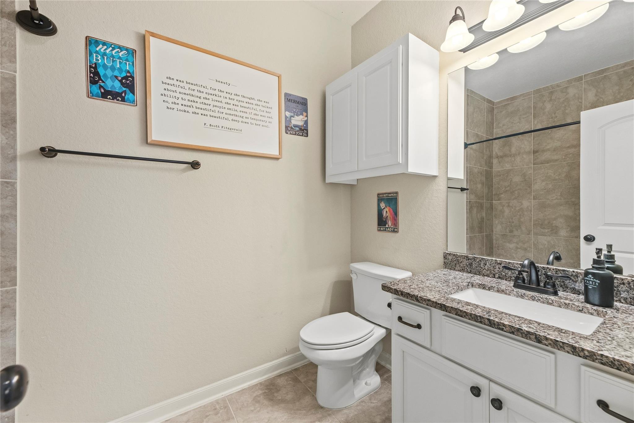 Bathroom with a granite vanity, white cabinetry, tiled shower, and playful framed wall art.