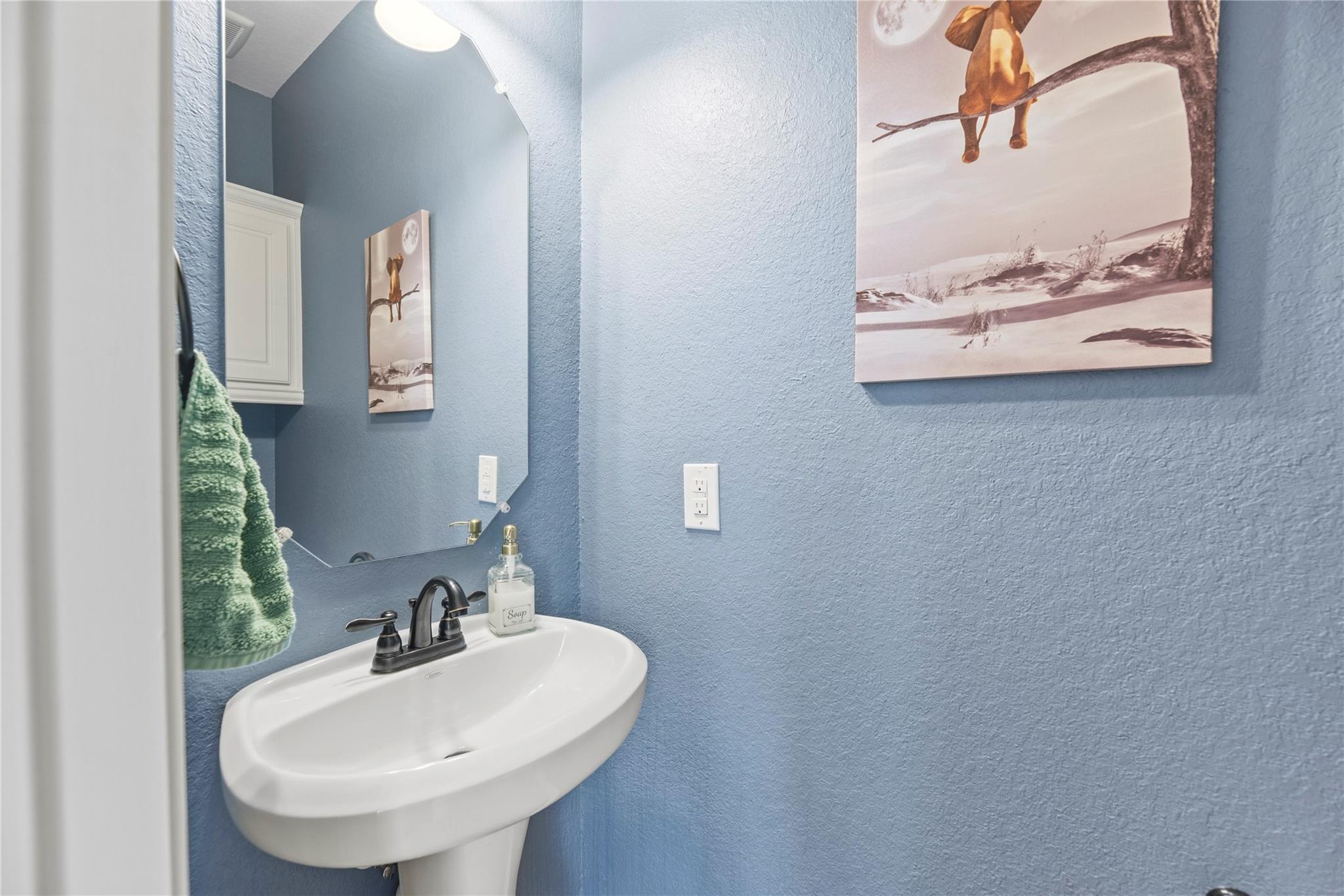 Bathroom pedestal sink with dark faucet against blue walls, beside a mirror and green hand towel.