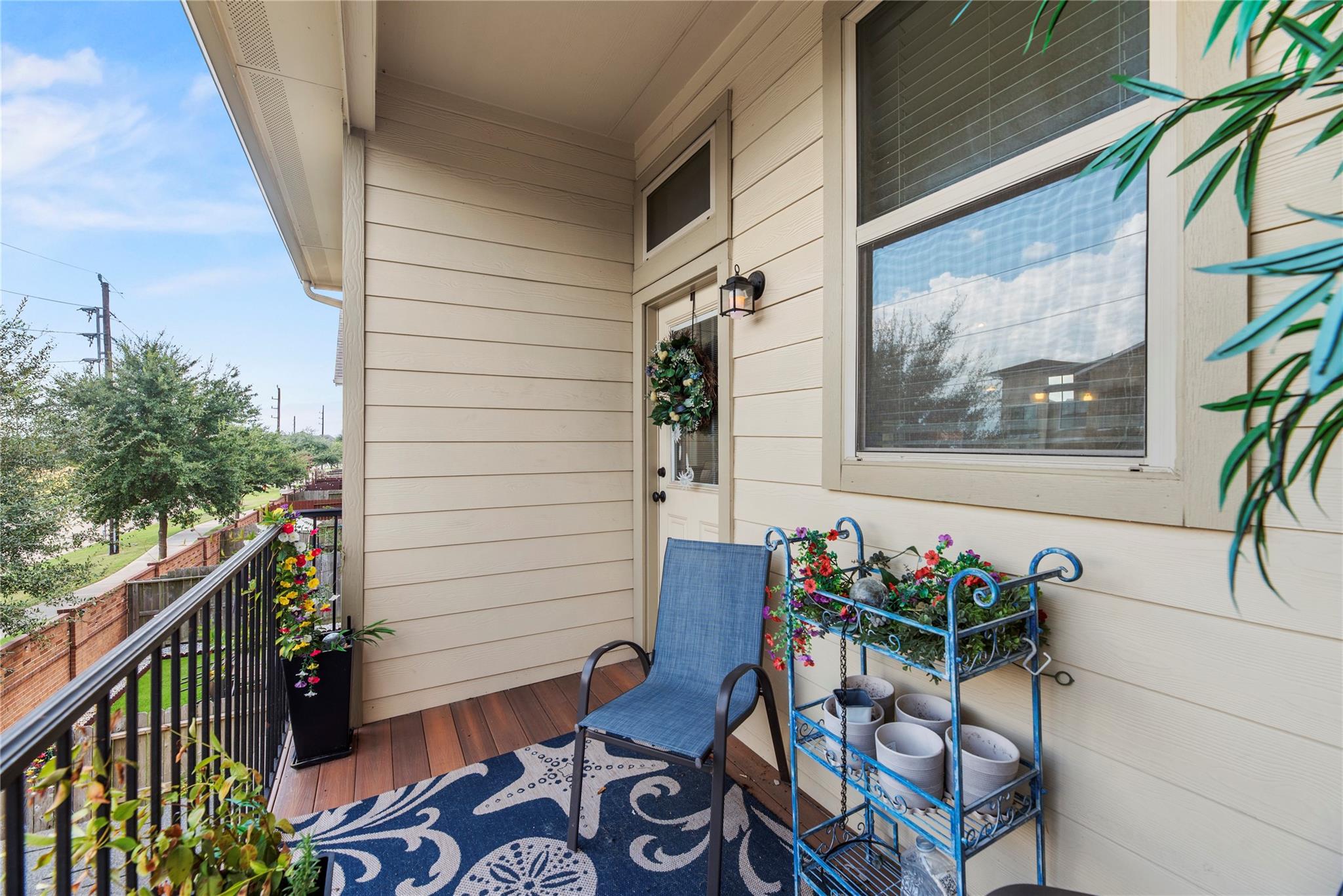 Furnished balcony with a blue chair, patterned rug, flower-filled planters, and views of trees and the street.