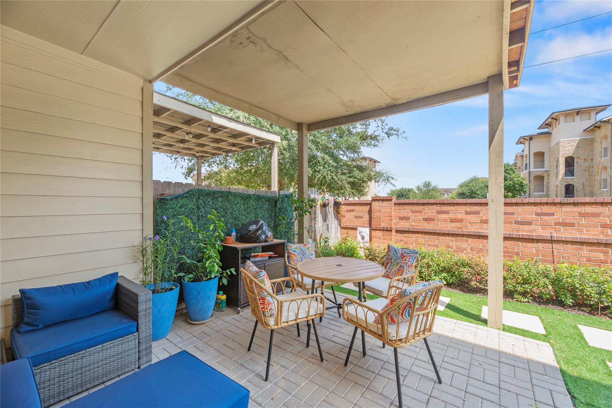 Covered patio with wicker dining chairs, blue-cushioned seating, potted plants, and a brick wall.