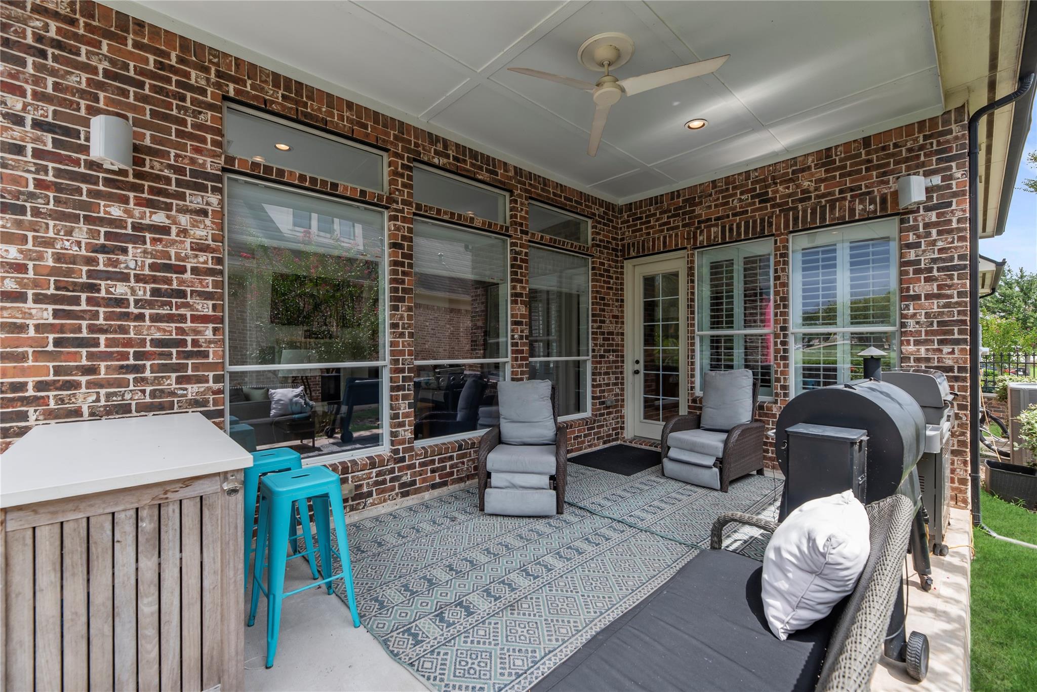 Covered brick patio with cushioned seating, patterned rug, ceiling fan, and outdoor grill.