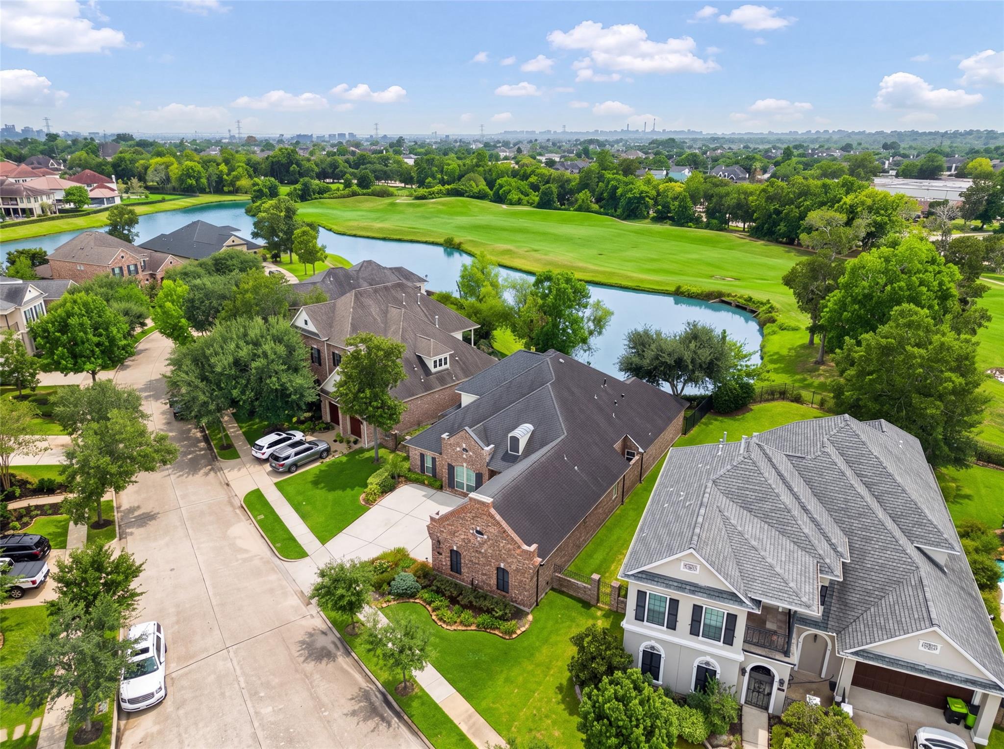 Aerial view of a brick home beside a pond and green golf course in a tree-lined neighborhood.