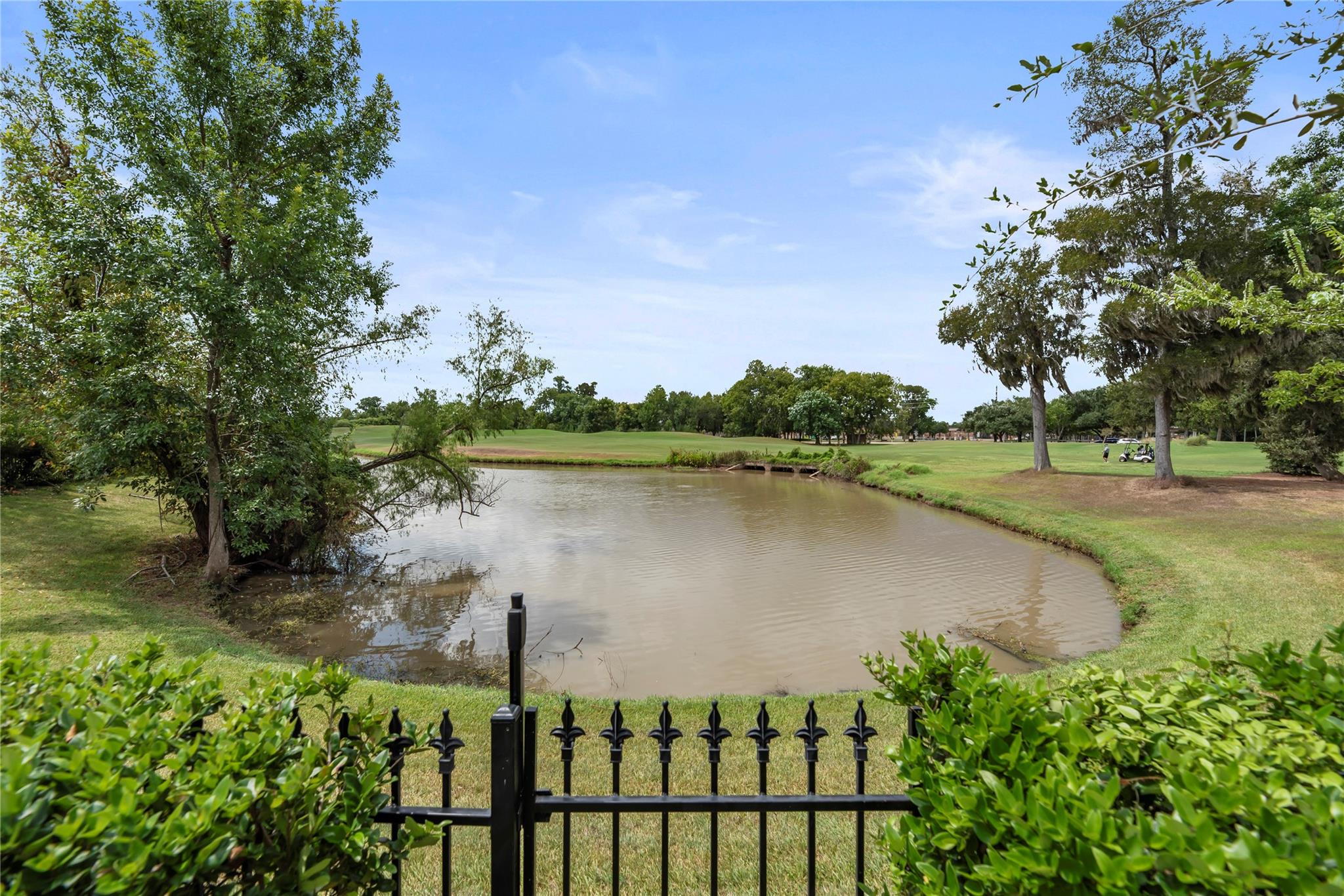Pond and golf course beyond a black ornamental fence, with mature trees and a golf cart in the distance.