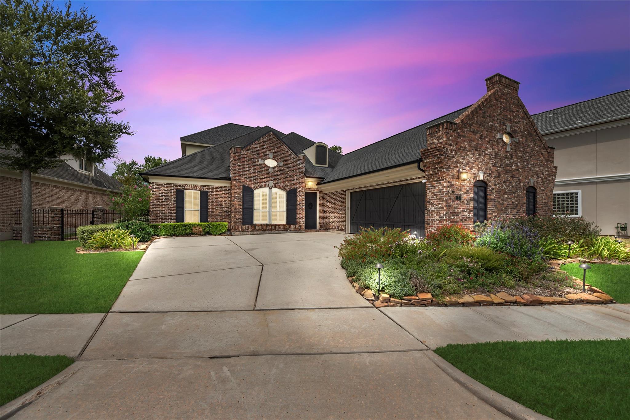 Red-brick home with a large attached garage, landscaped flower beds, and a broad driveway at dusk.