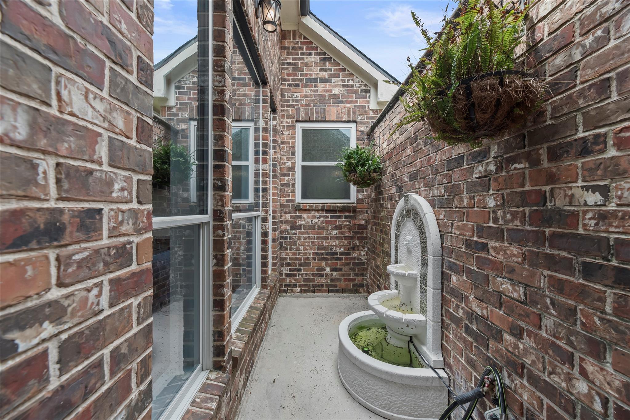 Brick courtyard with a white tiered fountain and hanging ferns beside tall windows.