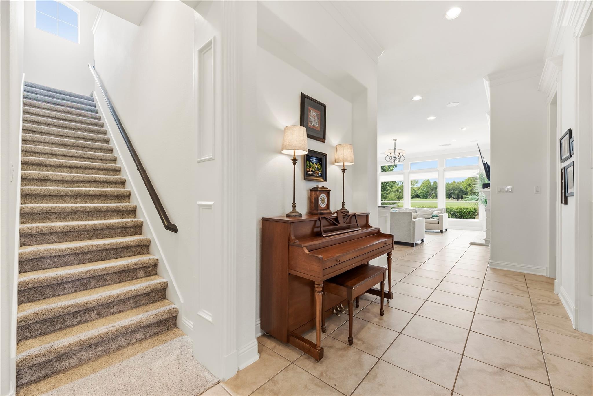 Foyer with a carpeted staircase, upright piano, and tiled floor leading to a sunny sitting area.