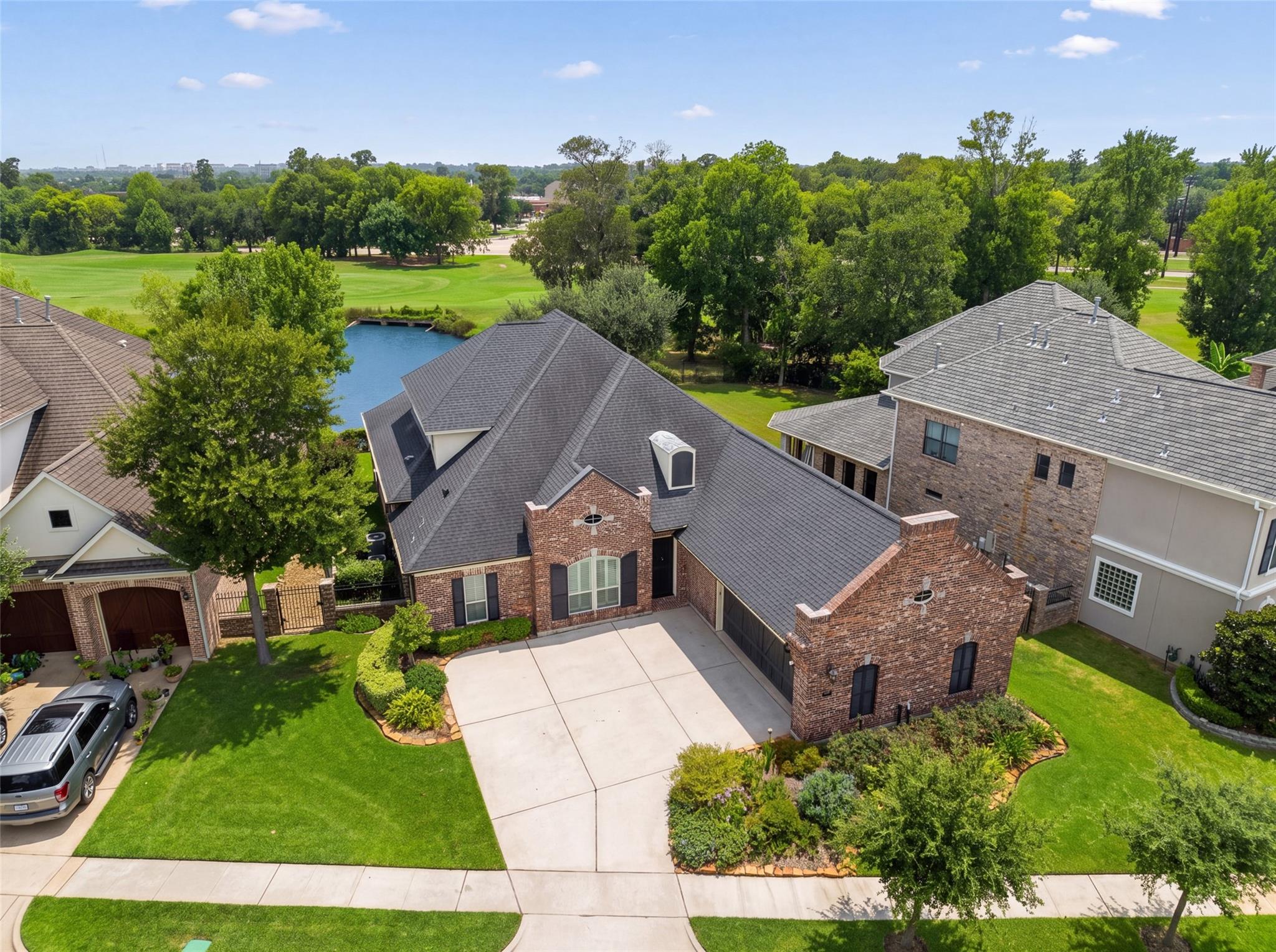 Aerial view of a brick home with a broad driveway, landscaped yard, and golf course and pond behind it.