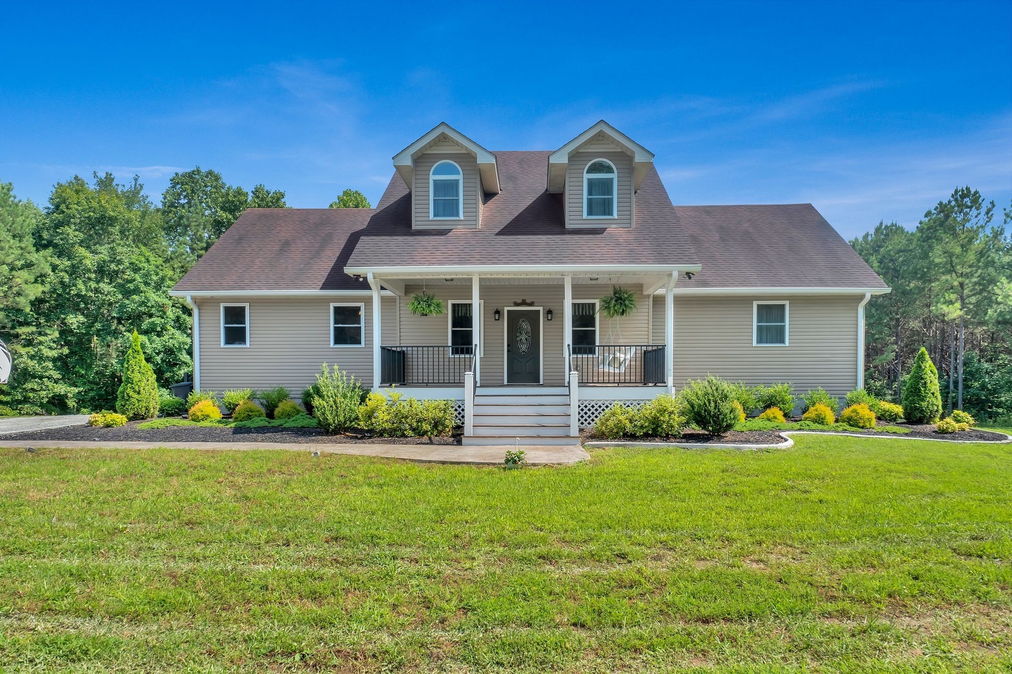Beige house with twin dormers, a covered porch, and landscaped front lawn.