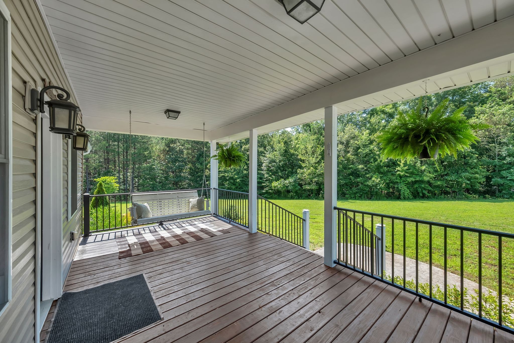 Covered wooden porch with a hanging swing and ferns overlooking a lawn and wooded yard.