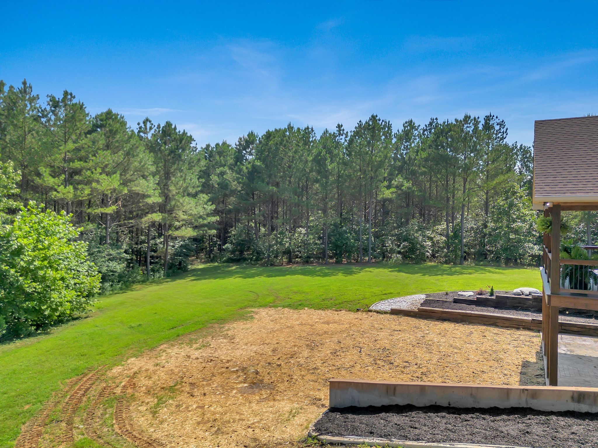 Backyard with a green lawn, wooded tree line, mulched landscaping, and part of a covered deck.