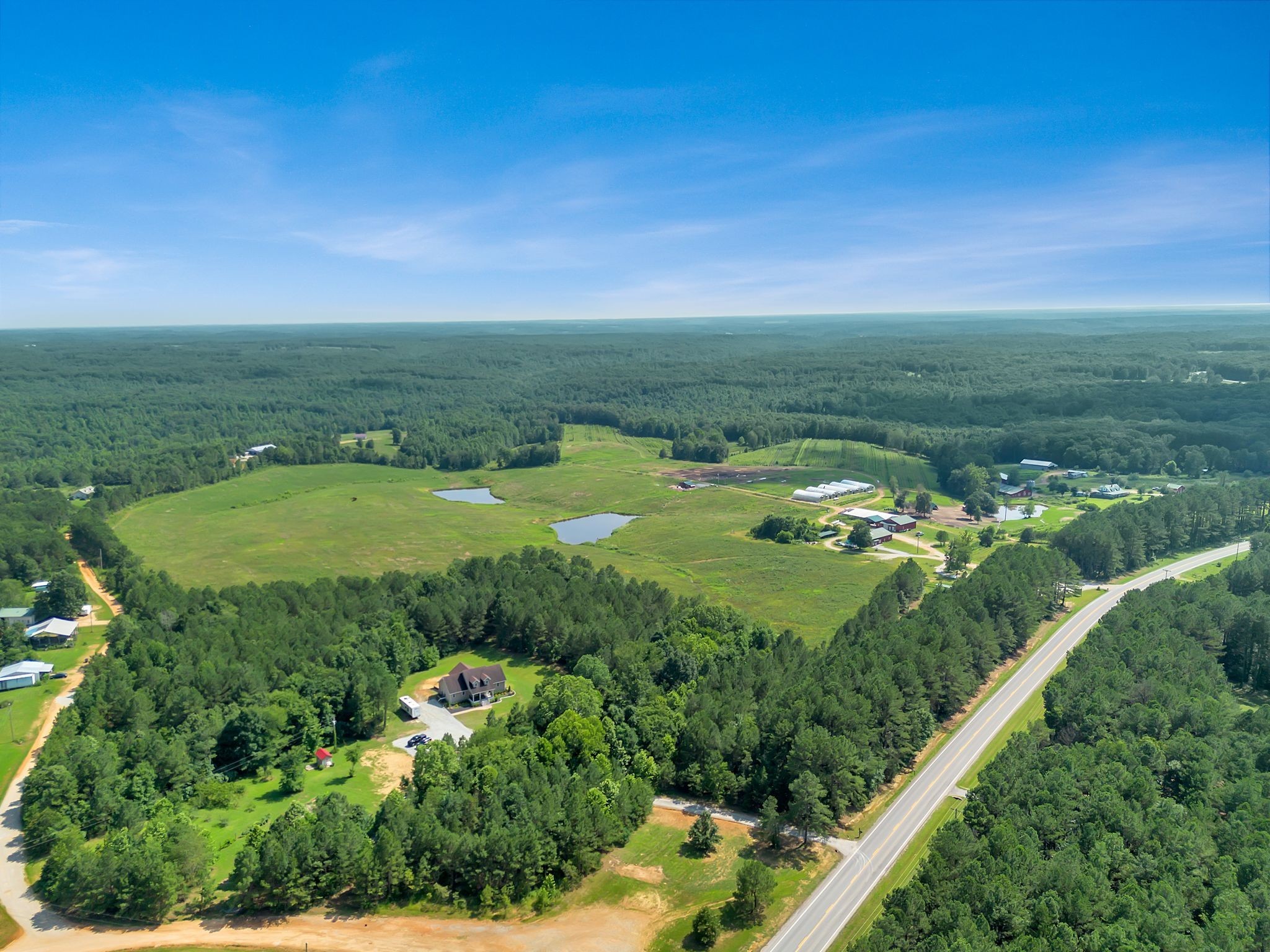 Aerial view of wooded acreage with a house, open fields, ponds, and farm buildings beside a highway.