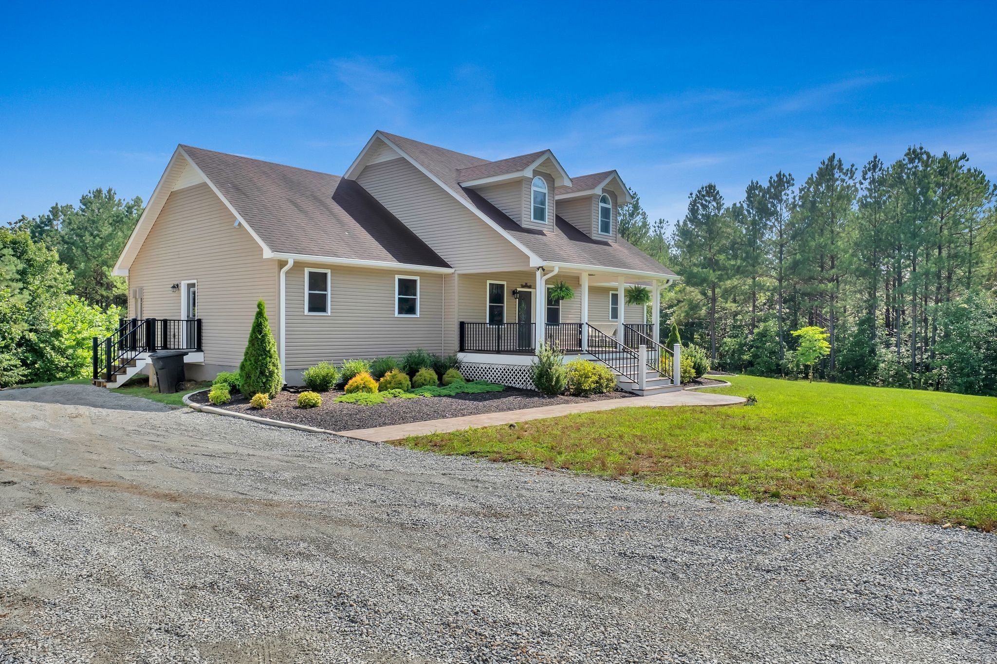 Beige-sided house with a covered porch and dormer windows beside a gravel driveway and pine trees.