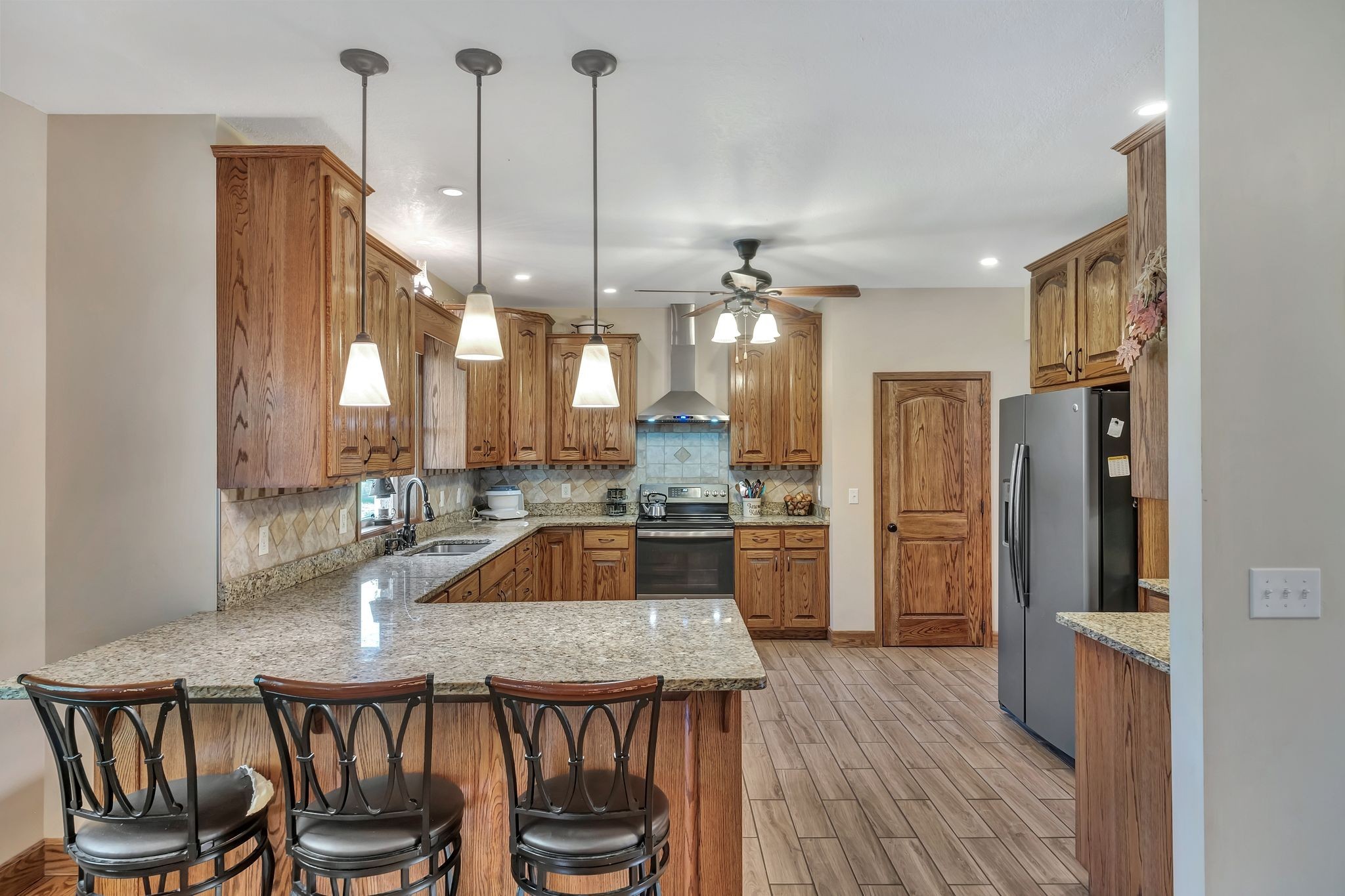 Kitchen with oak cabinetry, granite island, three bar stools, and stainless steel appliances.