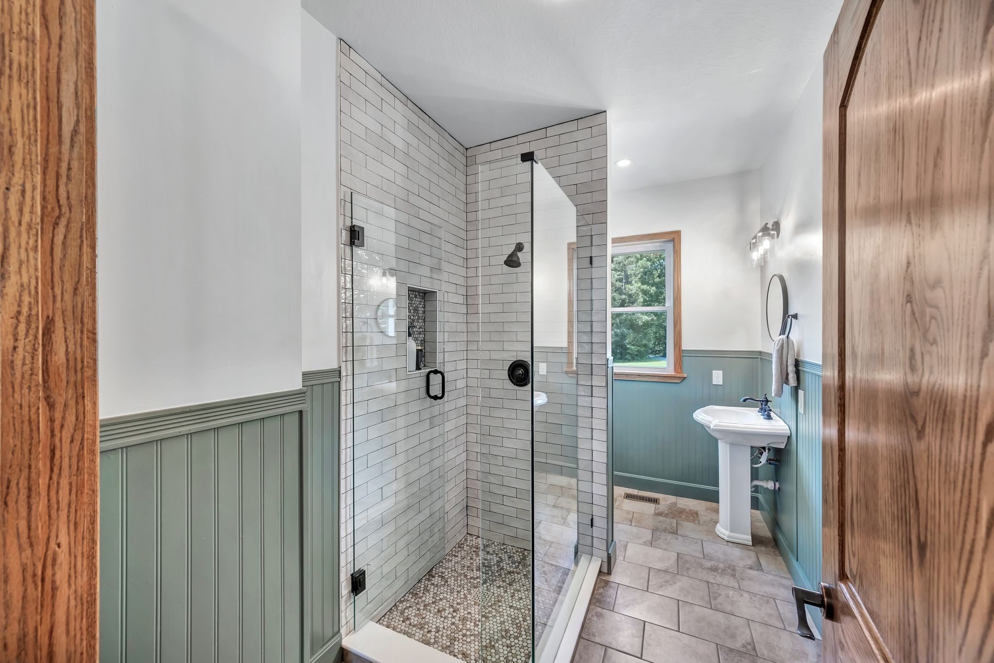 Bathroom with a glass subway-tile shower, sage-green wainscoting, and a pedestal sink by a wood-trimmed window.