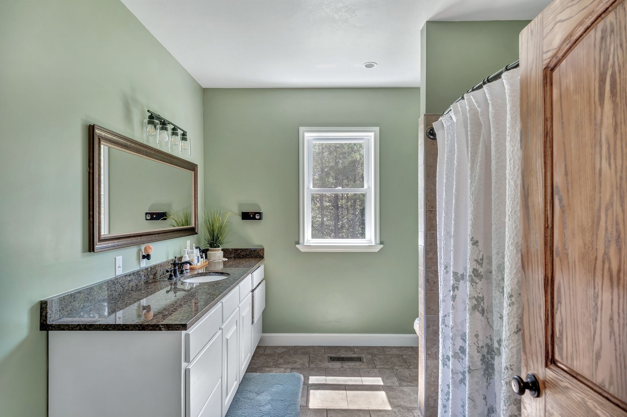Bathroom with a granite-topped vanity, pale green walls, and a tiled shower with a patterned curtain.